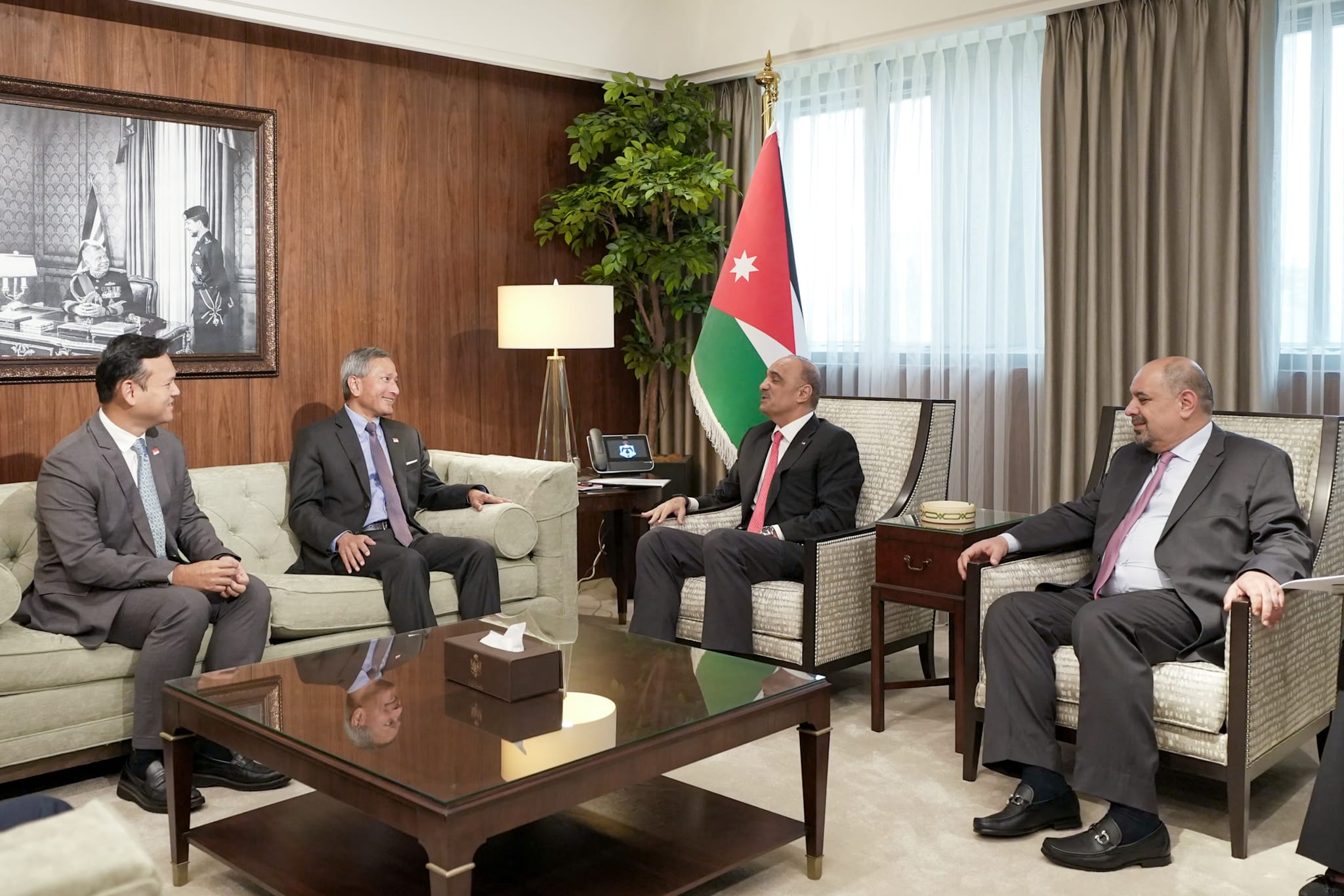Four men in suits sit in a room with a Jordanian flag and black and white portrait.